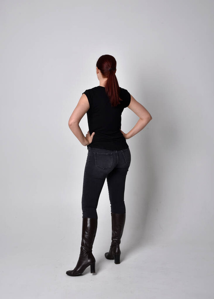 ポニーテールに赤い髪の女性のシンプルな全長の肖像画、カジュアルな黒のTシャツとジーンズを身に着けています。カメラに背を向けて立ちポーズ、スタジオの背景を背景に. - 写真・画像