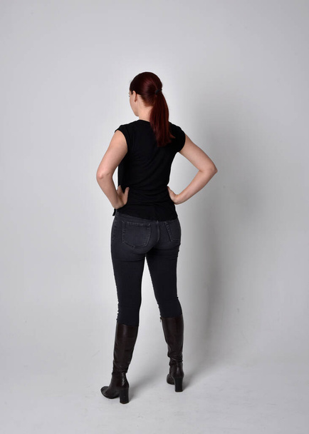 Retrato simple de mujer con el pelo rojo en una cola de caballo, con camiseta negra casual y jeans. Posar de pie con la espalda a la cámara de la cámara, sobre un fondo de estudio. - Foto, imagen
