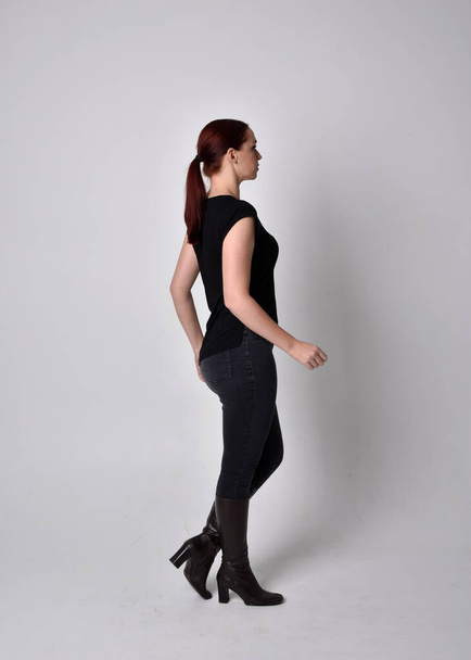 Simple full length portrait of woman with red hair in a ponytail, wearing casual black tshirt and jeans. Standing pose with back to the camera the camera, against a  studio background. - Foto, imagen