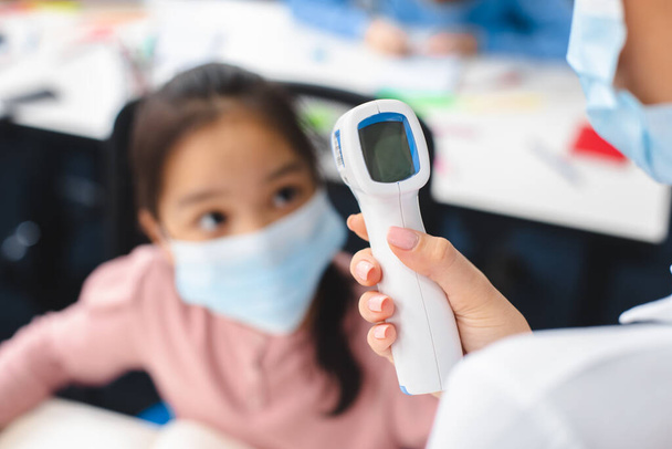 Учитель с бесконтактным термометром, измеряющим температуру азиатской девушки - Фото, изображение