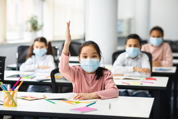 Kinderen steken hun hand op in de klas, dragen een medisch masker - Foto, afbeelding