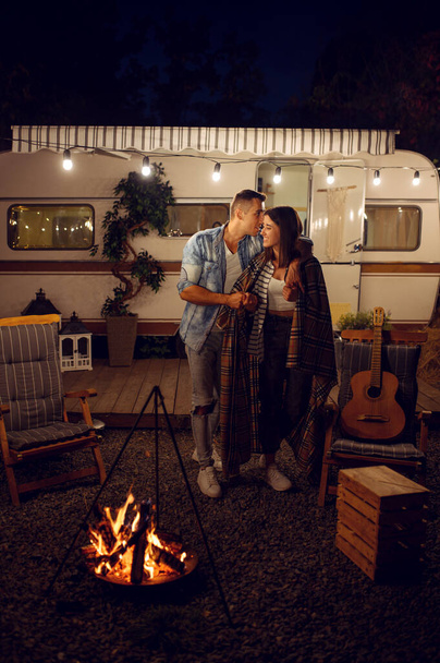 Jeune couple amoureux embrassant près du feu de camp, pique-nique au camping dans la forêt. Aventure estivale en VR, camping-car en arrière-plan. Loisirs de l'homme et de la femme près du feu de joie, voyageant avec remorque - Photo, image