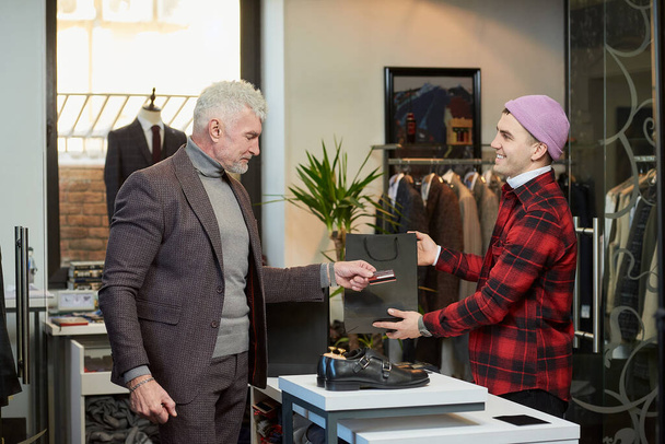 Un hombre maduro con el pelo gris y un físico deportivo está entregando una tarjeta de crédito a un vendedor para pagar una compra en una tienda de ropa. Un asistente de tienda con una sonrisa está dando una bolsa de papel a un cliente masculino - Foto, imagen