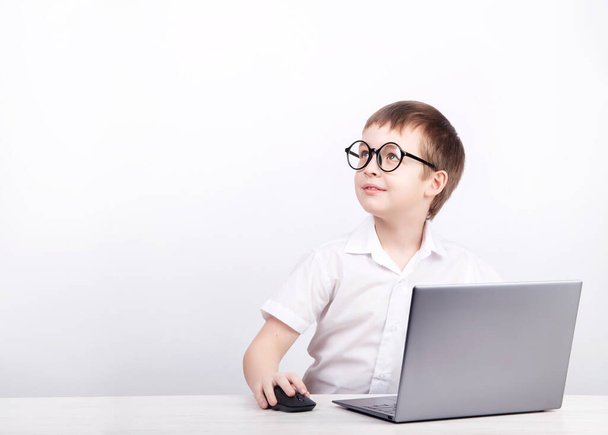 chlapec s brýlemi, student základní školy, sedí u stolu s laptopem na bílém pozadí - Fotografie, Obrázek