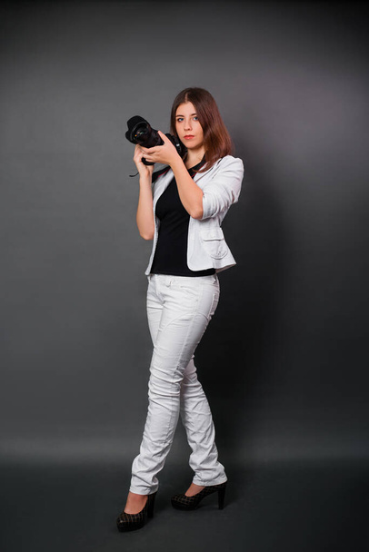 黒い背景に立っている間、カメラを手に白いスーツを着た女性がポーズをとっている。スタジオの現代写真家. - 写真・画像