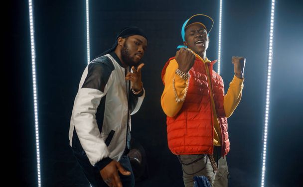 Twee stijlvolle rappers poseert in gloeiende kubus, studio met donkere achtergrond. Hiphopartiesten, breakdansers - Foto, afbeelding