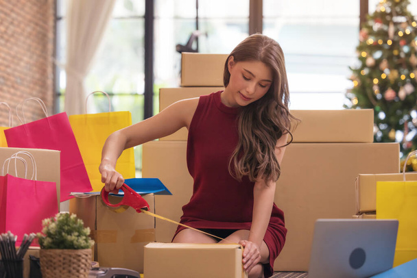 Empresaria encintando una caja de cartón, mujer asiática empacando cajas entre una pila de paquetes en su negocio en línea de compras en casa - Foto, imagen