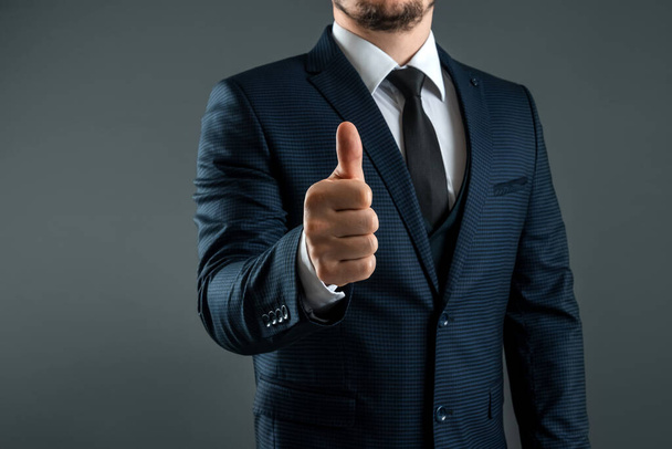 Male hand in suit shows thumbs up gesture on gray background. Concept ok, approval, close-up - Фото, зображення