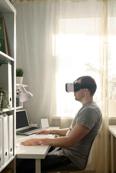 Genç adam yurt içi ofisinde arttırılmış VR kulaklık kullanıyor. VR kulaklıklı bir erkek. Ev hayatı, pencereden yansıyan doğal ışık. - Fotoğraf, Görsel