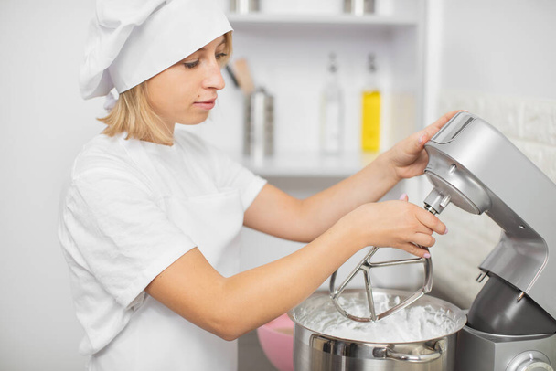 Pretty young blond girl confectioner, using modern electric food processor to mixing the dough for baking a cake or cookies in light kitchen interior - Foto, Bild