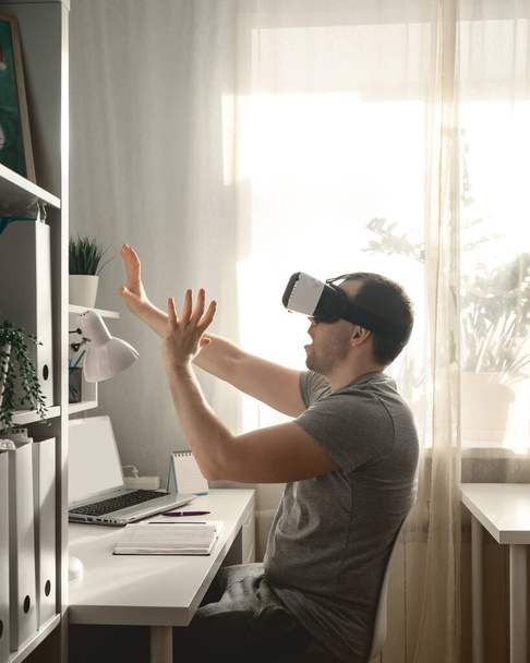 Egy fiatalember, aki kibővített VR fejhallgatót használ az irodában egy lezárás alatt Covid-19 miatt. Oldalsó nézet Férfi VR headsetben. Családi élet, természetes háttérvilágítás az ablakból - Fotó, kép