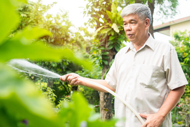 A happy and smiling Asian old elderly man is watering plants and flowers for a hobby after retirement in a home. Concept of a happy lifestyle and good health for seniors. - Photo, Image