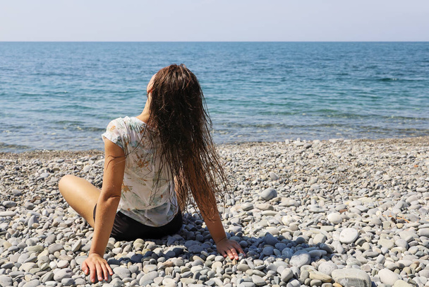 Genç bir kadın sırt üstü çakıl taşı plajında tek başına oturur ve ıslak saçlarını sallar. - Fotoğraf, Görsel
