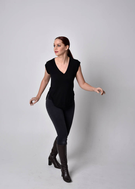 Simple full length portrait of woman with red hair in a ponytail, wearing casual black tshirt and jeans. Standing pose front on with hand gestures, against a  studio background. - Foto, imagen