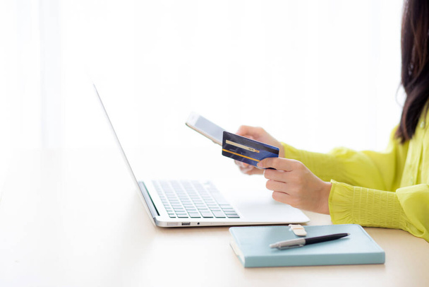 Крупный план молодых азиатских деловых женщин, использующих смартфон и держащих кредитные карты во время онлайн-покупок и оплаты с ноутбуком на столе дома, женщина держит дебетовую карту, коммуникационная концепция. - Фото, изображение