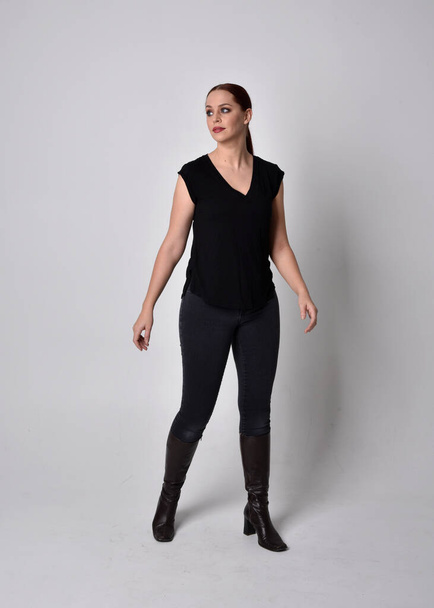 Simple full length portrait of woman with red hair in a ponytail, wearing casual black tshirt and jeans. Standing pose facing front on, against a  studio background. - Photo, Image