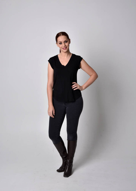 Simple full length portrait of woman with red hair in a ponytail, wearing casual black tshirt and jeans. Standing pose facing front on, against a  studio background. - Фото, изображение