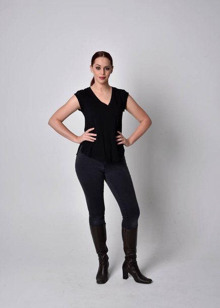 Simple full length portrait of woman with red hair in a ponytail, wearing casual black tshirt and jeans. Standing pose facing front on, against a  studio background. - Fotografie, Obrázek