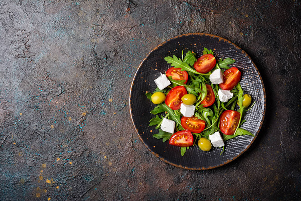 Top view of healthy and tasty vegetarian food. Salad with fresh arugula leaves, cherry tomatoes, soft cheese and green olives on dark concrete background with copy space - Photo, image