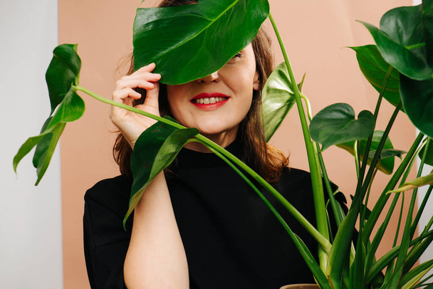 Sorrindo mulher de meia idade espreitando de trás planta monstera - Foto, Imagem