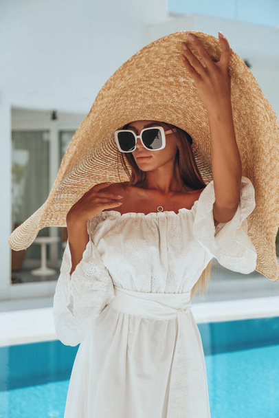 fashion holiday photo of beautiful sexy woman with dark hair in elegant dress and big hat posing in beach club with swimming pool  - Valokuva, kuva