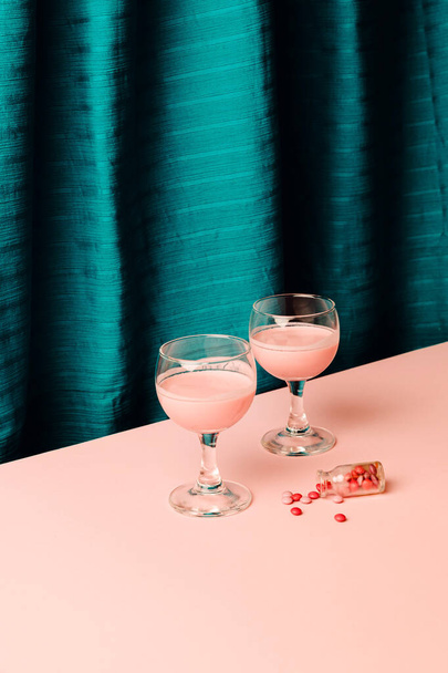 Dois copos de vinho com bebida criativa e pílulas parecem um doce parecido ou suor colocar em um chão com pequena garrafa contra fundo rosa e tecido de seda ciano azul. - Foto, Imagem