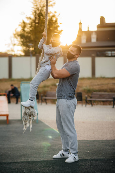 A father helps his son climb a rope on a sports field in masks during sunset. Healthy parenting and healthy lifestyle - Foto, afbeelding