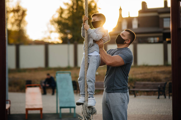 A father helps his son climb a rope on a sports field in masks during sunset. Healthy parenting and healthy lifestyle - Fotó, kép
