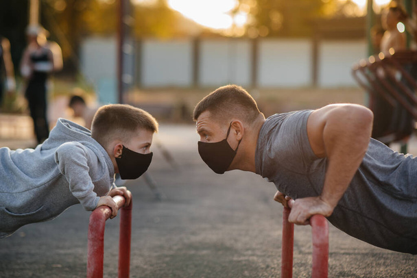 Πατέρας και γιος παίζουν σπορ στο γήπεδο φορώντας μάσκες κατά τη διάρκεια του ηλιοβασιλέματος. Υγιής γονική μέριμνα και υγιεινός τρόπος ζωής - Φωτογραφία, εικόνα