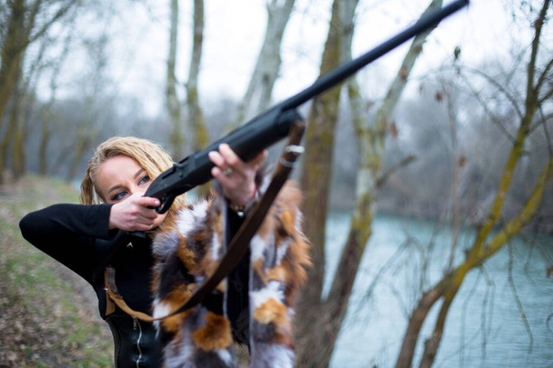 Kaukasische vrouw met blauwe ogen in zwart pak en vacht op de schouder mikken met een jachtgeweer in het voorste deel bij een rivier - Foto, afbeelding