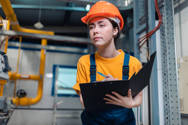 Industrial production. Portrait of a young woman in uniform and helmet with a folder and a pen in her hand, writing meter readings. In the background-boiler equipment and pipes. Bottom view. - 写真・画像