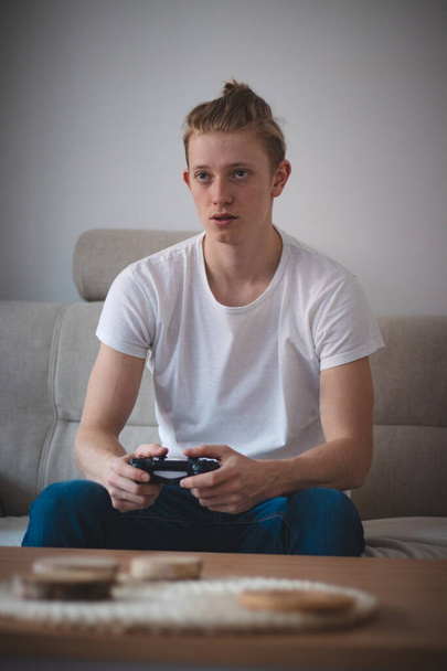 Soustředěný výraz muže ve věku 20-29 let s blonďatými vlasy. Hráč sedí na gauči v obývacím pokoji a hraje na herní konzoli. Dnešní svět e-sporťáků. Nuda v karanténě. - Fotografie, Obrázek