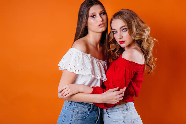 Zwei junge und schöne Mädchen zeigen Emotionen und lächeln im Studio auf orangefarbenem Hintergrund. Mädchen für die Werbung - Foto, Bild