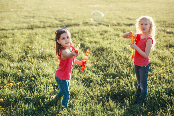 Happy Caucasian girls blowing soap bubbles in park on summer day. Kids having fun outdoor. Authentic happy childhood magic moment. Lifestyle seasonal activity for children. - Photo, Image