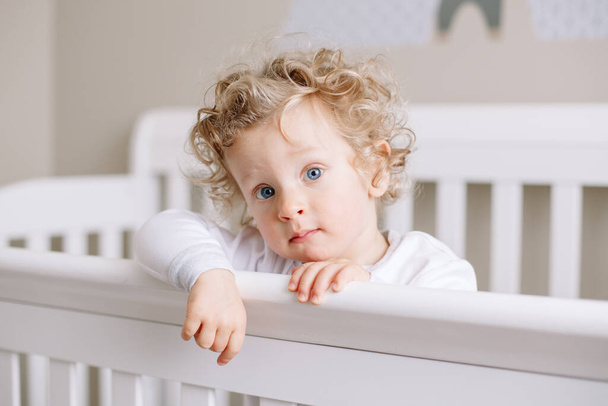Cute adorable baby boy toddler standing in crib at kids nursery room at home. Curious charming baby boy with curly blond hair and blue eyes looking at camera. Happy authentic candid home life.  - Fotografie, Obrázek