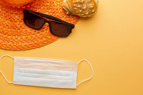 Γυαλιά ηλίου και καπέλο σε πορτοκαλί φόντο με προστατευτική μάσκα προσώπου, έννοια της πανδημίας διακοπές στην παραλία. - Φωτογραφία, εικόνα