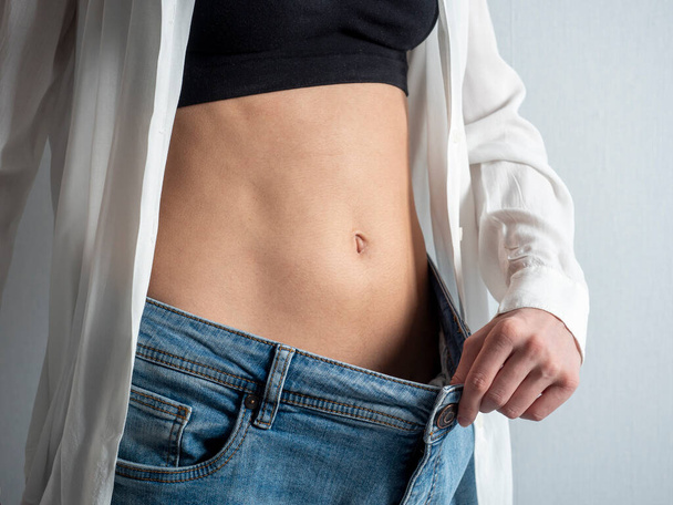 струнка жінка з голим шлунком показує, як вона схудла, тримаючи джинси рукою. Концепція дієти, схуднення, здоровий спосіб життя
 - Фото, зображення