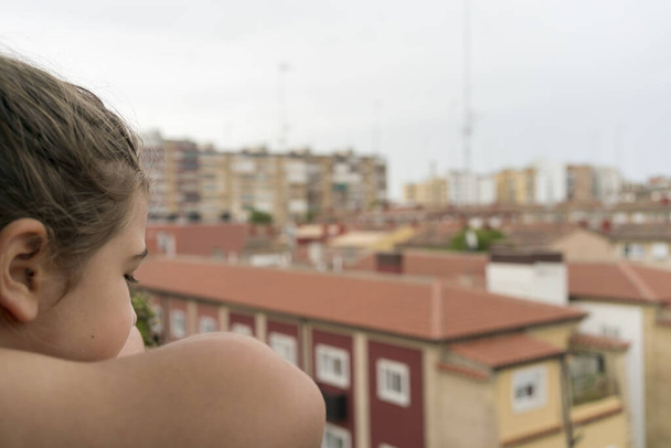 Teras çıkıntısına yaslanmış bir kız, Covid-19 'un hapsedildiği sokağı izliyor. Sosyal uzaklık kavramı. - Fotoğraf, Görsel
