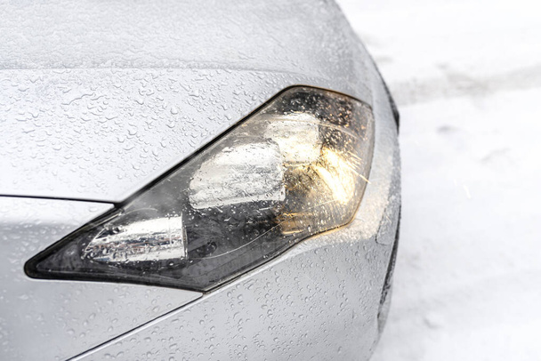 De gedompelde koplampen op een zilveren auto op een besneeuwde weg in het bos, zichtbare sneeuw die valt. - Foto, afbeelding