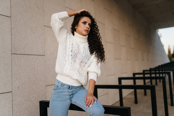 Beautifully posing photographer brunette in jeans and a long sweater, lifting his hand with his curly hair - Fotografie, Obrázek