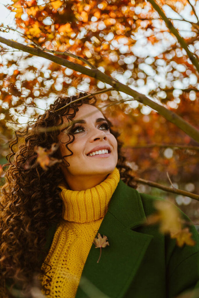 Bright beautiful brunette with curly curly hair and smooth white teeth opened in her charming smile looks a burning look in the sky against the background of yellow autumn leaves and harmonizes with - Fotoğraf, Görsel