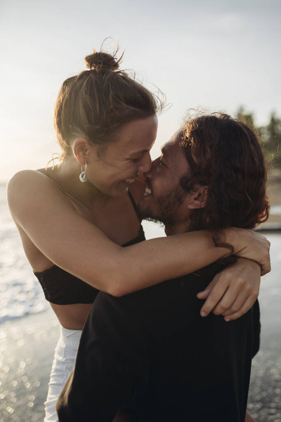 A bright couple in love with a man lifted the girl as she wrapped her arms around his neck their looks joined while smiles are adorable against the ocean - Photo, image