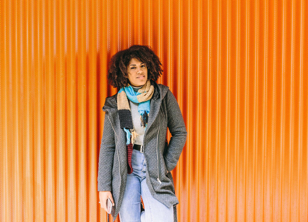 Chica de estilo afroamericano con abrigo gris y jeans frente a una pared naranja - Foto, Imagen