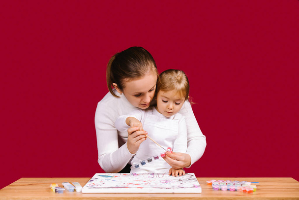 Home family activity. Mom help little daughter to paint with brush and acrylic kit a picture on numbers. Curious blonde little girl is excited of workshop - Foto, Bild