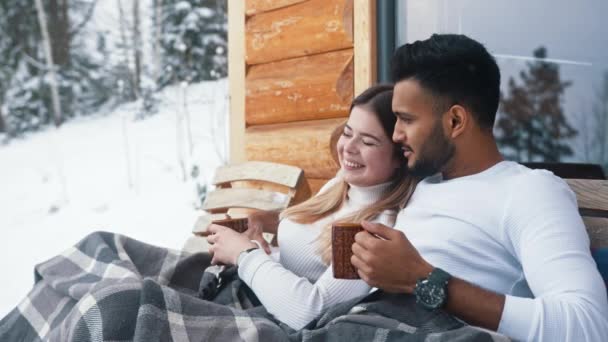 Feliz joven pareja abrazándose cubierta con manta en el banco al aire libre. Beber bebidas calientes y disfrutar de la nieve - Metraje, vídeo