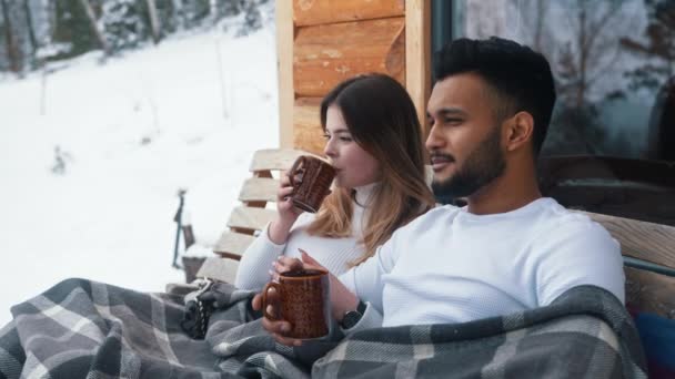 Joyeux jeune couple étreignant couvert de couverture sur le banc extérieur. Boire une boisson chaude et profiter de la neige - Séquence, vidéo