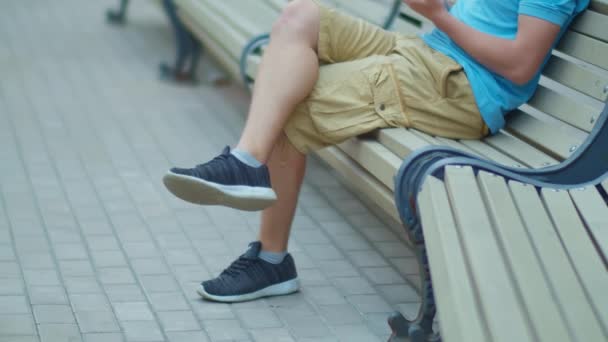 Πόδια του ανθρώπου σε σορτς κάθεται στο παγκάκι της πόλης με κινητό τηλέφωνο παρακολούθησης κάμερα - Πλάνα, βίντεο