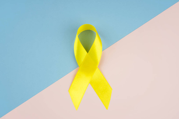 Κίτρινη κορδέλα ευαισθητοποίησης για τον καρκίνο ως σύμβολο της ευαισθητοποίησης για τον παιδικό καρκίνο σε μπλε και ροζ φόντο. Αντιγραφή χώρου - Φωτογραφία, εικόνα