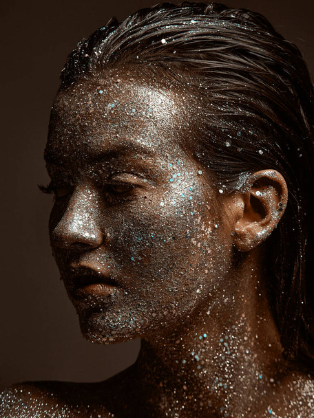 Silver sparkling female face, young girl posing with festive glowing glittering make up close up studio portrait - 写真・画像