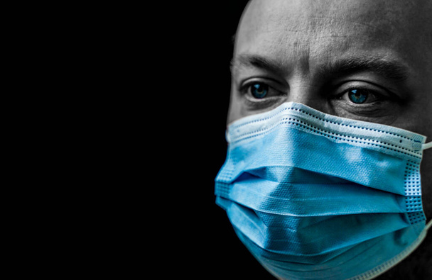 Sick man with bright blue eyes wearing a facemask, portrait isolated on black with shallow depth of field. High quality photo - Photo, Image